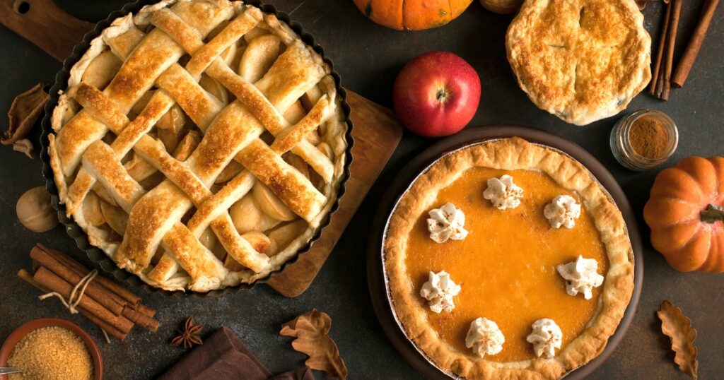 A festive, autumn-inspired display of pumpkin and apple pies, surrounded by cinnamon sticks, apples, leaves and small gourds.