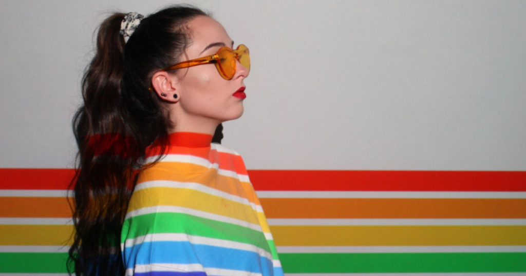 A woman wearing a ponytail with large, orange sunglasses and a rainbow top stands against a rainbow wall to support Pride Month.